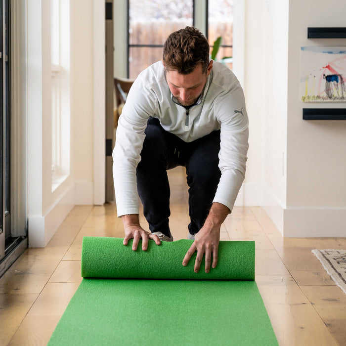 A man in a white jacket kneels on a wooden floor, unrolling a green exercise mat—ideal for indoor golf practice—next to the BirdieBall Zippered Storage Bag, available in various sizes, in a bright hallway.