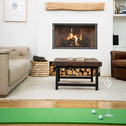 A living room with a lit fireplace, beige sofa, brown leather chair, wooden coffee table, basket of books, and a BirdieBall 2' x 10' Putting Mat + Stance Mat Package - Indoor RollTech™ Putting Green with two golf balls for at-home practice.