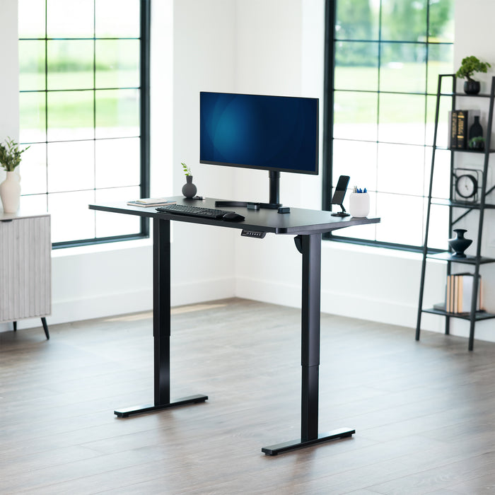 Black adjustable desk with a computer setup in a bright room with large windows.
