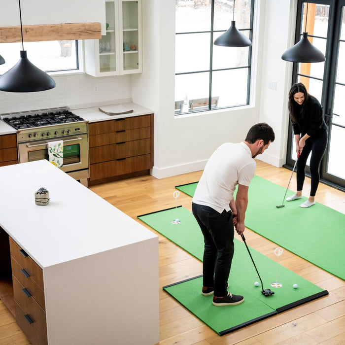 Two people play mini-golf on a BirdieBall Golf Stance Mat in a modern kitchen with wood floors, white walls, large windows, and stainless steel appliances.