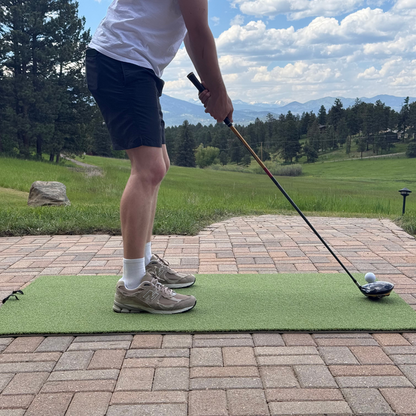 A golfer in shorts and sneakers readies a drive on the BirdieBall Premium Turf Golf Hitting Mat, set outdoors amid grass and trees.