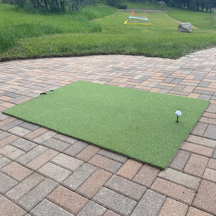 A golf ball on a tee rests on the BirdieBall Premium Turf Golf Hitting Mat, set up on a paved patio with lawn and equipment visible in the background.