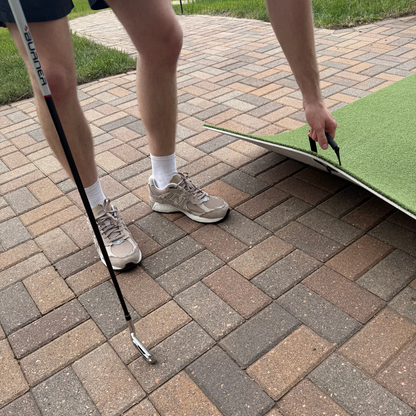 A person holds a golf club in one hand and lifts a BirdieBall Premium Turf Golf Hitting Mat made of green artificial turf on a brick patio.