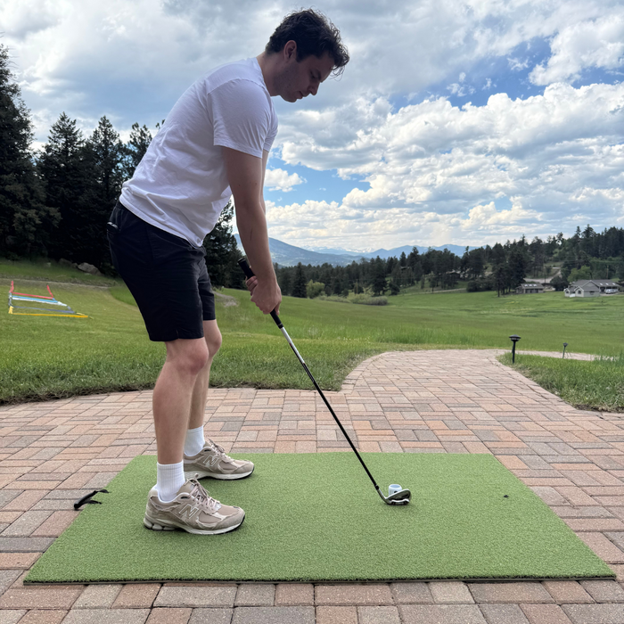 A golfer stands on the BirdieBall Premium Turf Golf Hitting Mat on a brick patio, club in hand, ready to swing at a ball with scenic trees and mountains in the background.