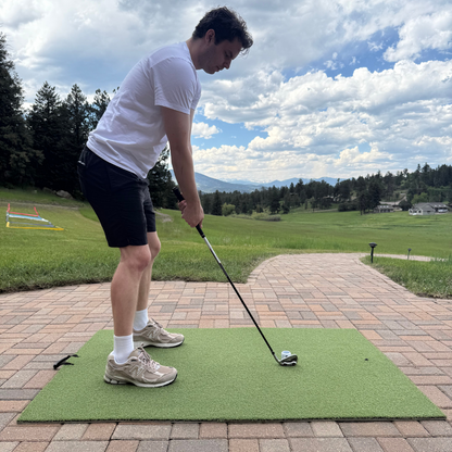A golfer stands on the BirdieBall Premium Turf Golf Hitting Mat on a brick patio, club in hand, ready to swing at a ball with scenic trees and mountains in the background.