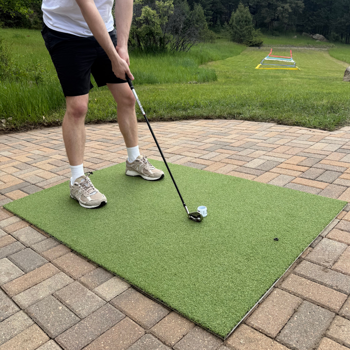 A person in shorts and sneakers prepares to hit a golf ball with a club on the BirdieBall Premium Turf Golf Hitting Mat, placed on a brick patio.
