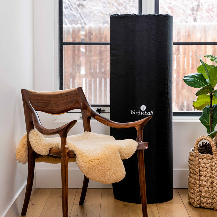 A wooden chair with a cream sheepskin sits next to a tall, black BirdieBall Zippered Storage Bag in a bright room with a window and potted plant—perfect for convenient indoor putting green storage.